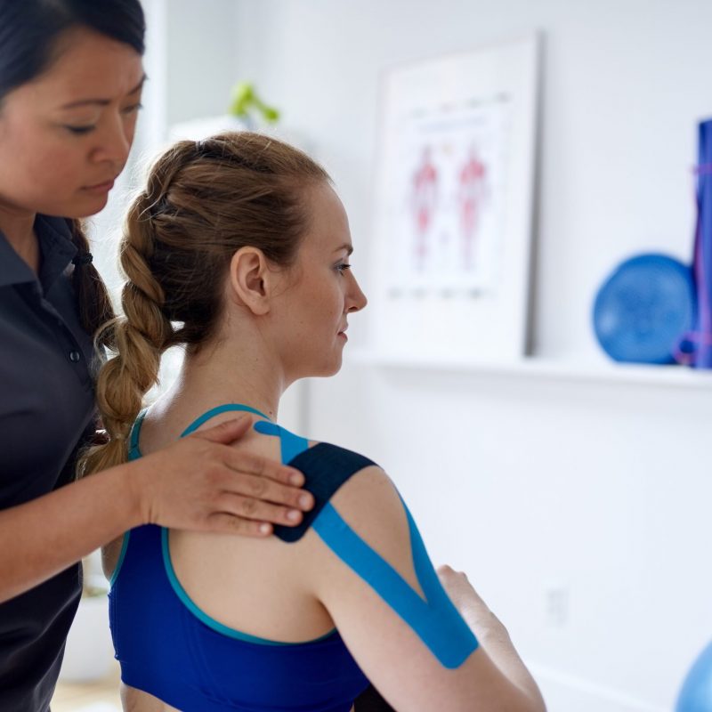 Candid image of an attractive patient during appointment with a professional asian physiotherapist, in design kinesiotherapy clinic stretching medical kinesio tape for neck and shoulder pain.
